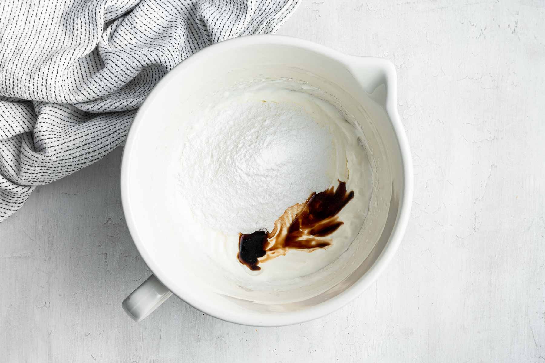 Adding vanilla and powdered sugar to freshly whipped cream in a white bowl.