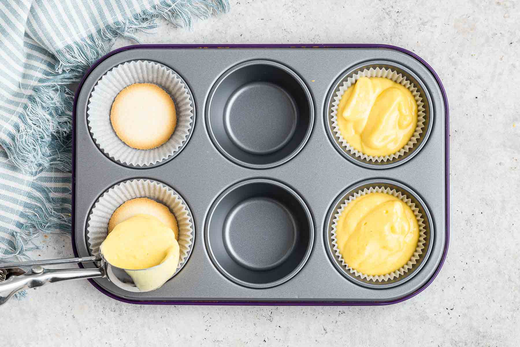 A silver cookie scoop dropping yellow batter in a muffin pan.