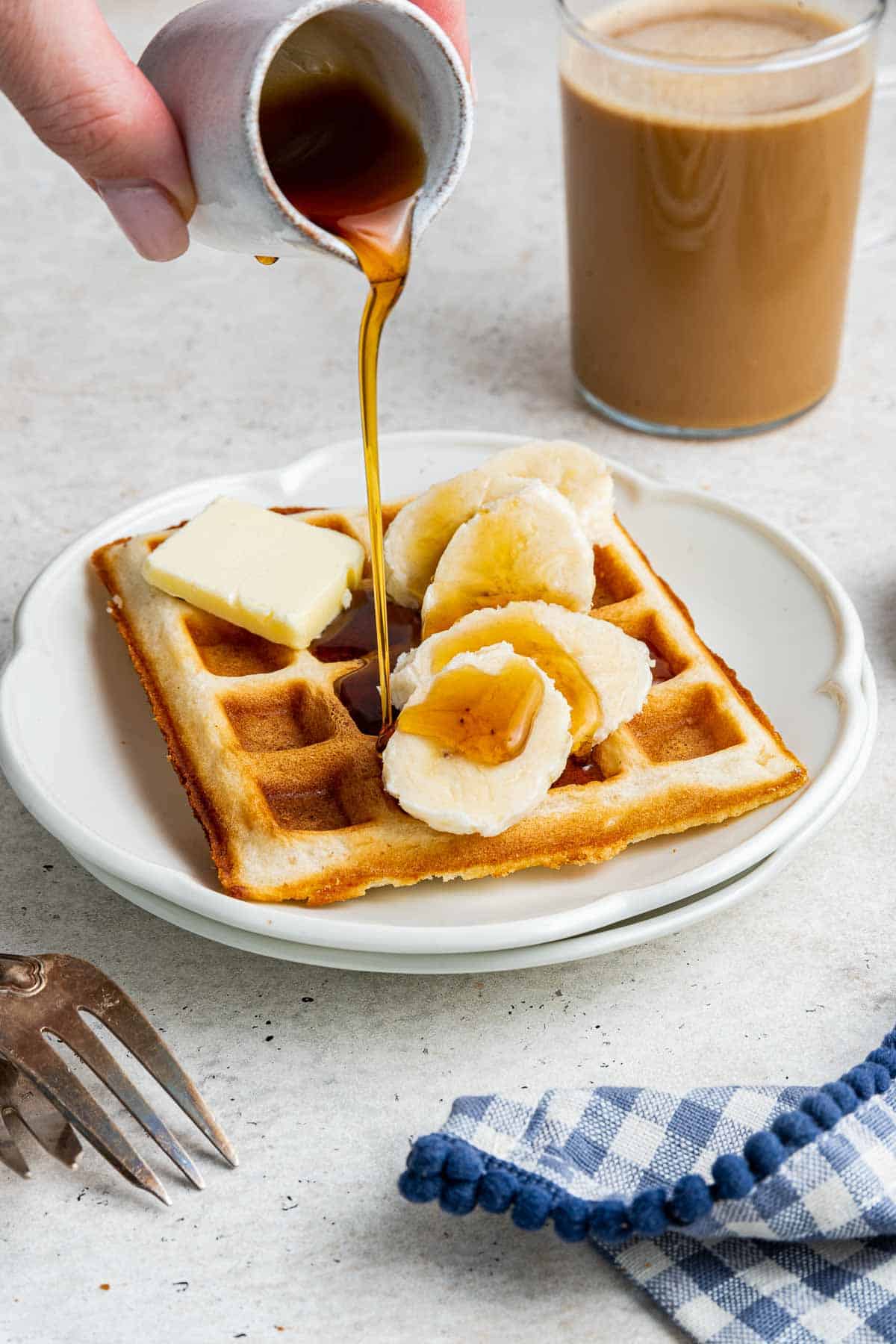 Handing pouring maple syrup on a single waffles for one on a round plate.