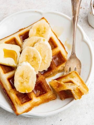 Single waffle topped with sliced banana, maple syrup with a fork on the side.