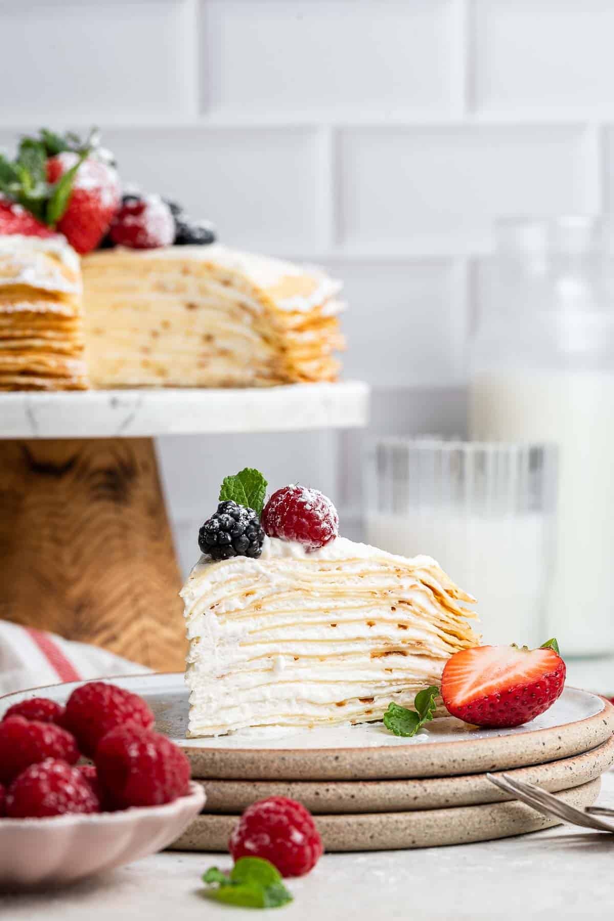 Crepe cake on a cake stand with one piece missing on plate and fresh berries.
