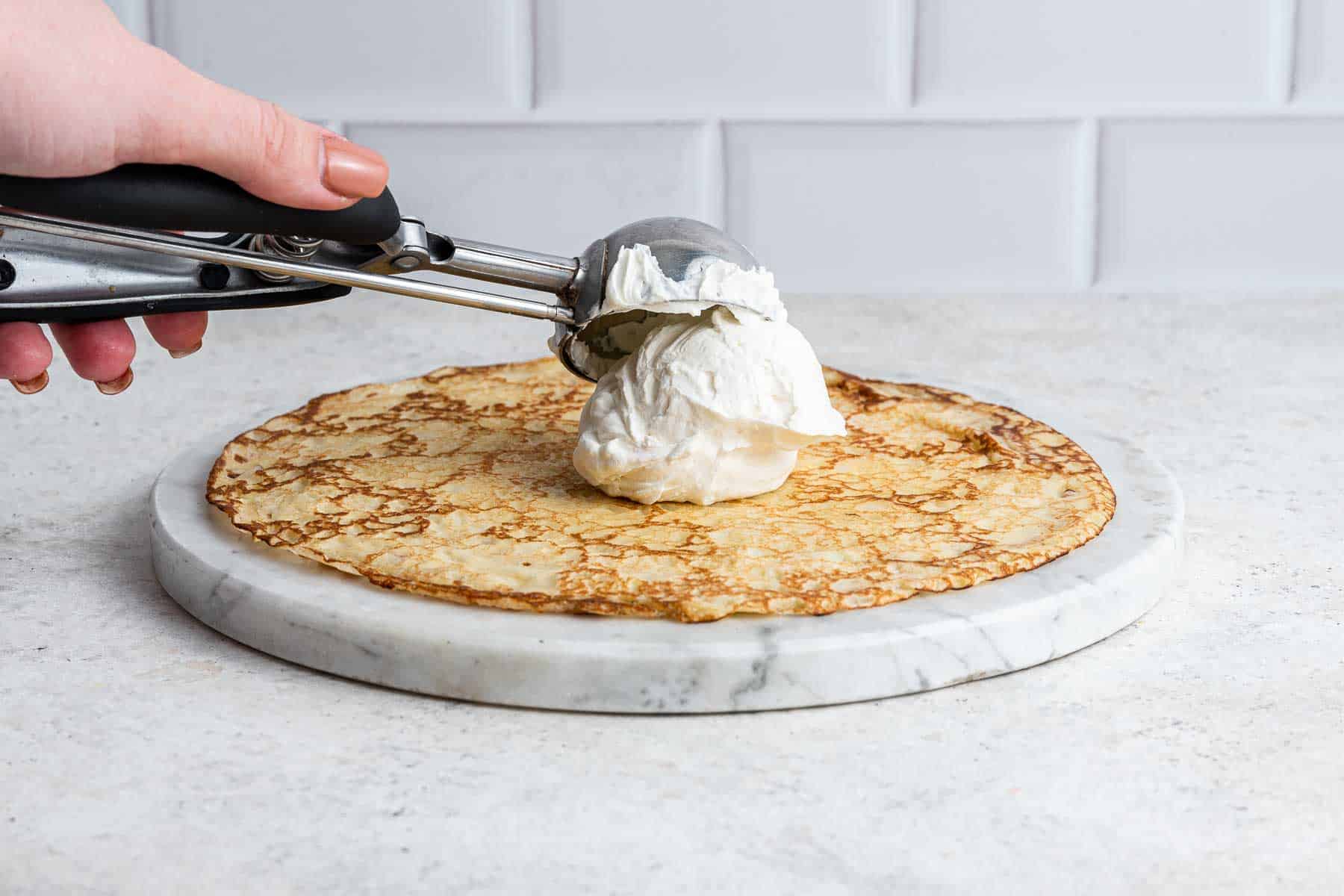 Placing mascarpone filling between layers of a crepe cake with ice cream scoop.