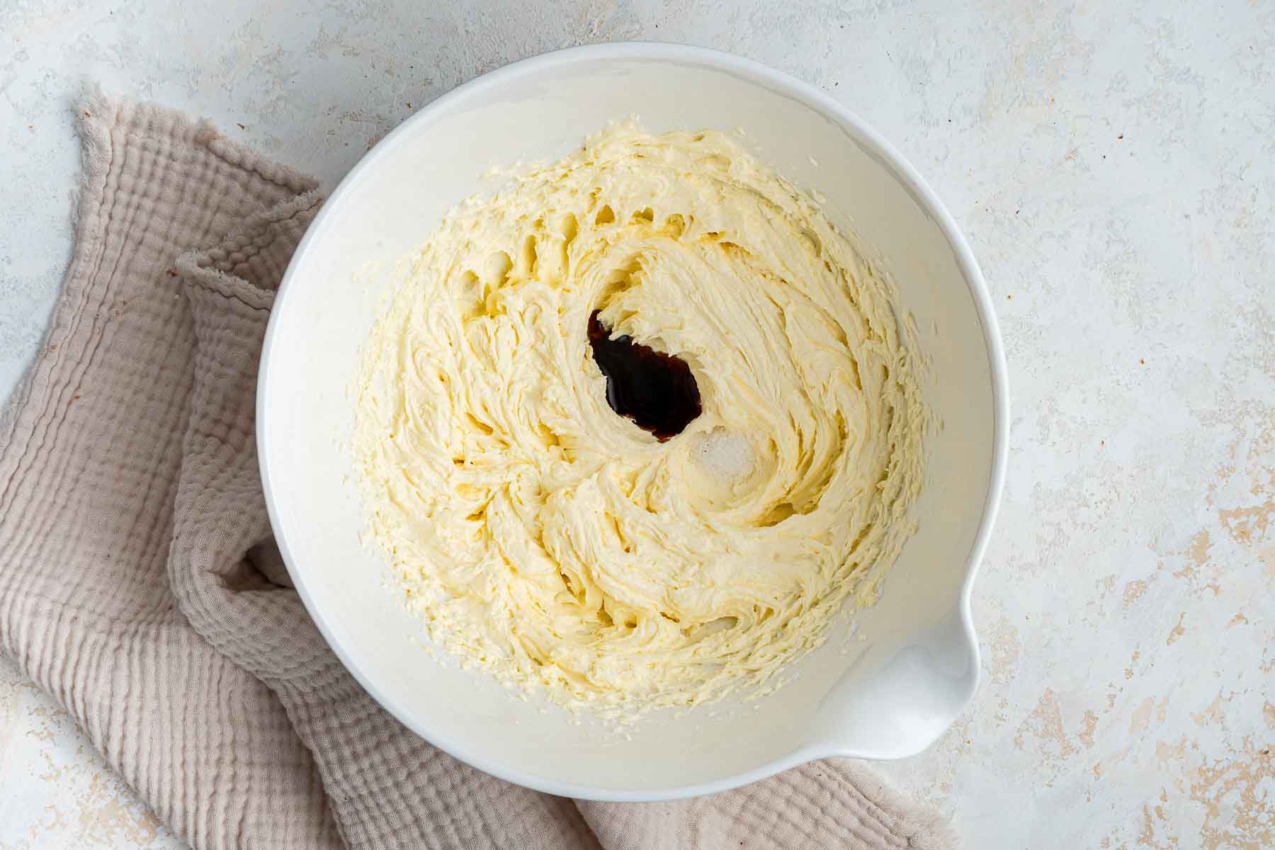 Drops of vanilla and salt in a yellow frosting in white mixing bowl.