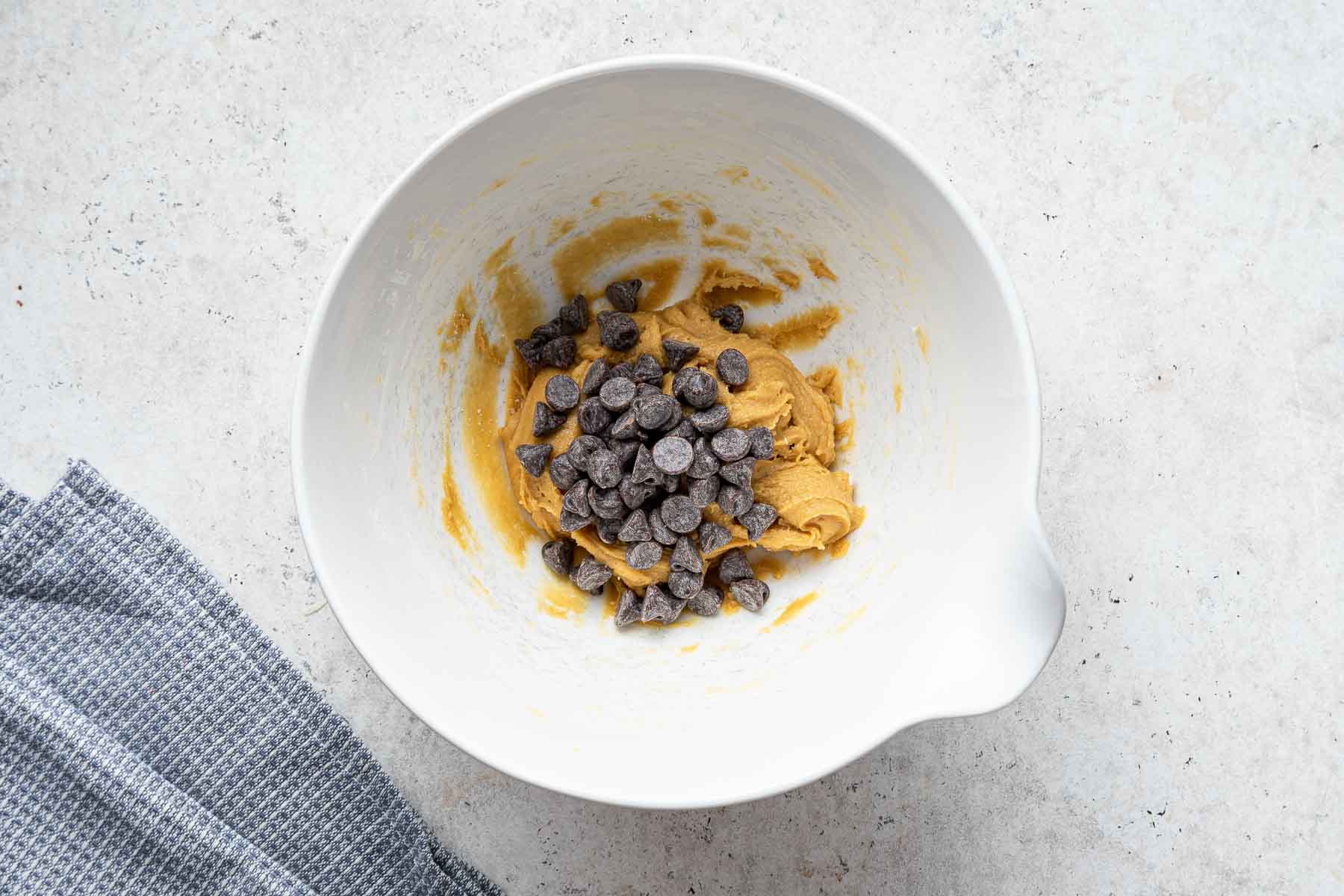 Adding chocolate chips to a single chocolate chip cookie recipe in white bowl.