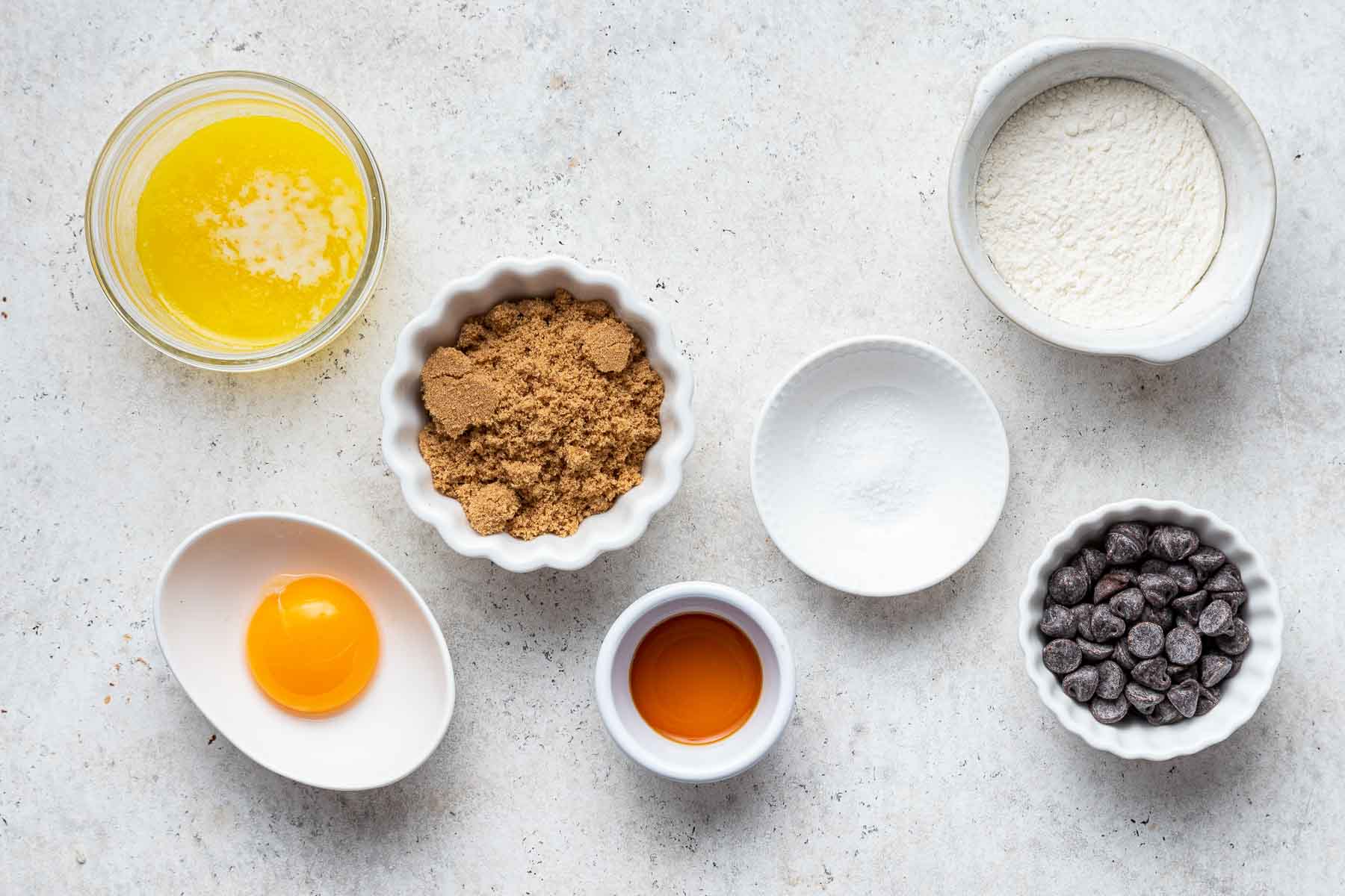 Small white bowls with melted butter, egg yolk, brown sugar, and chocolate chips on grey counter.