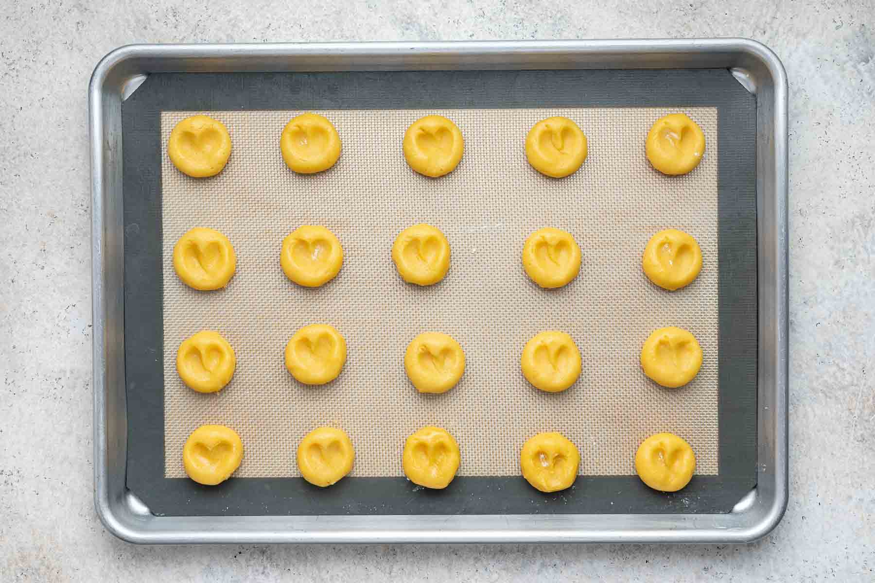 Twenty yellow cookie dough balls with heart shape indents on a cookie sheet.