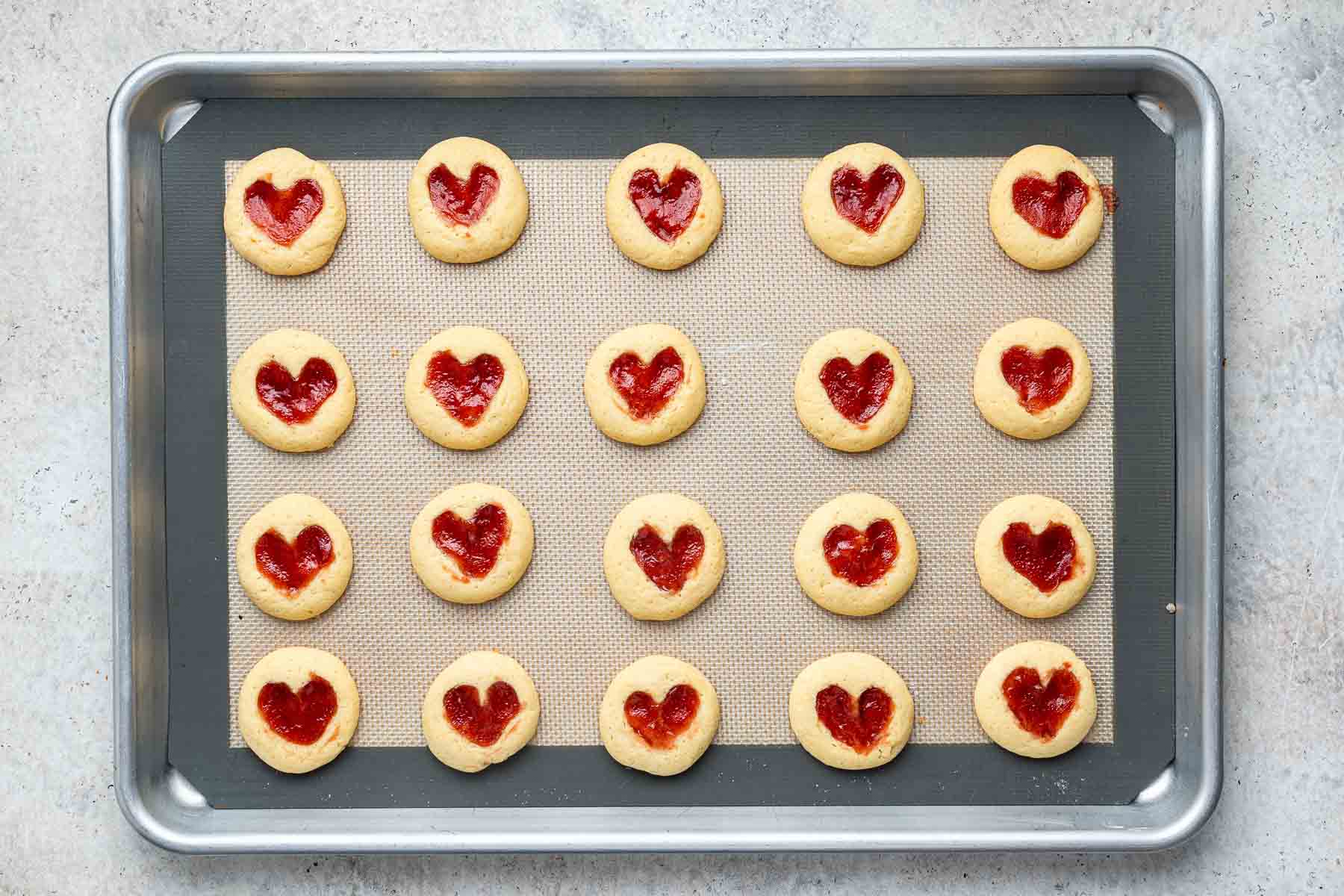 Strawberry jam cookies with hearts in the center on baking sheet.