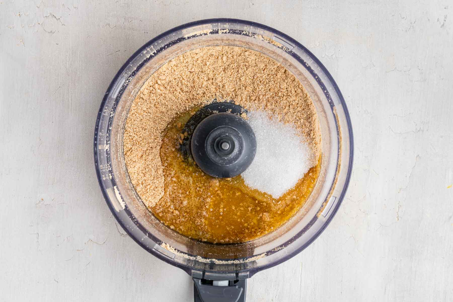 Small food processor bowl that contains graham cracker crumbs, melted butter and sugar.