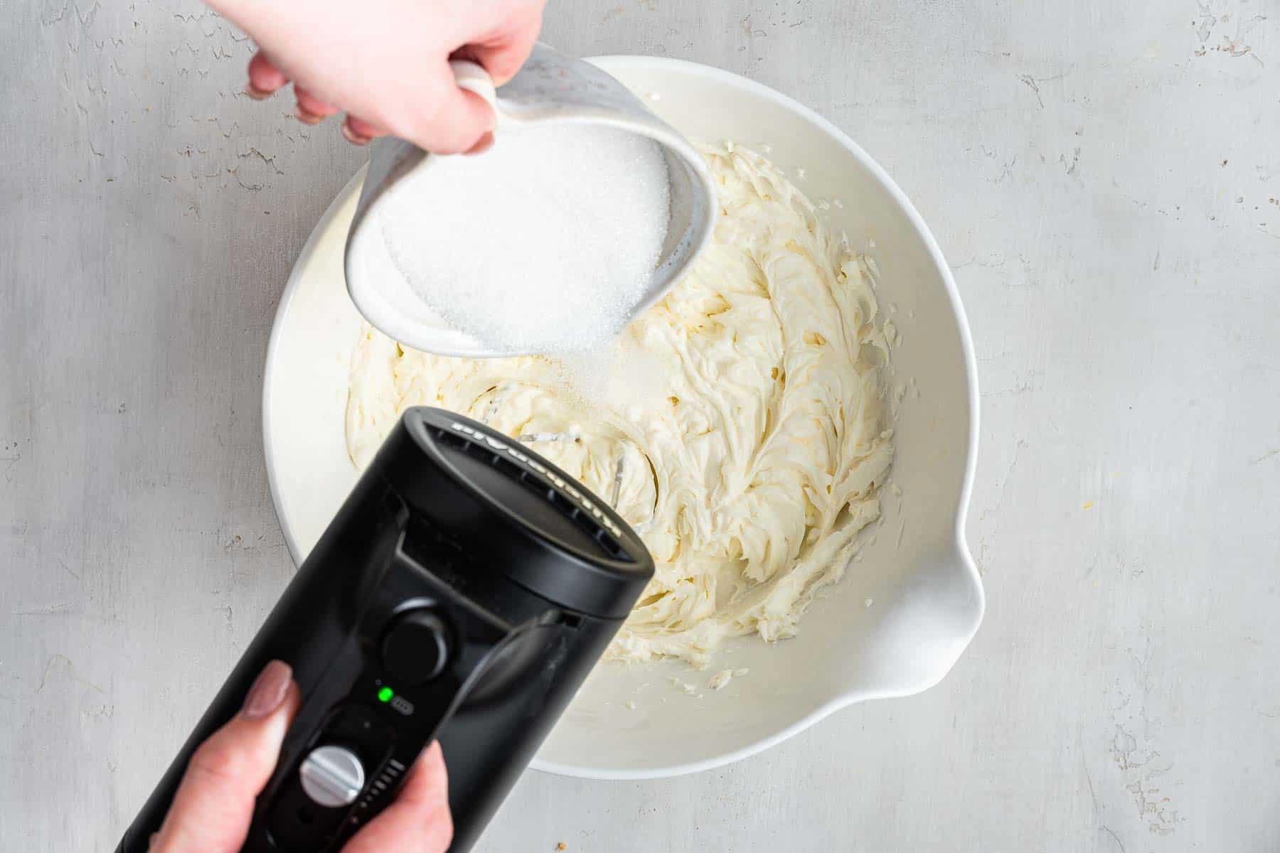 Bowl of white batter with sugar being poured in while mixing with electric mixer.