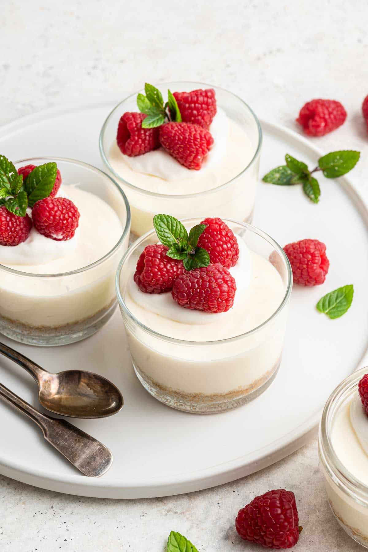 White plate with 3 no bake cheesecake cups on top, garnished with fresh raspberries.