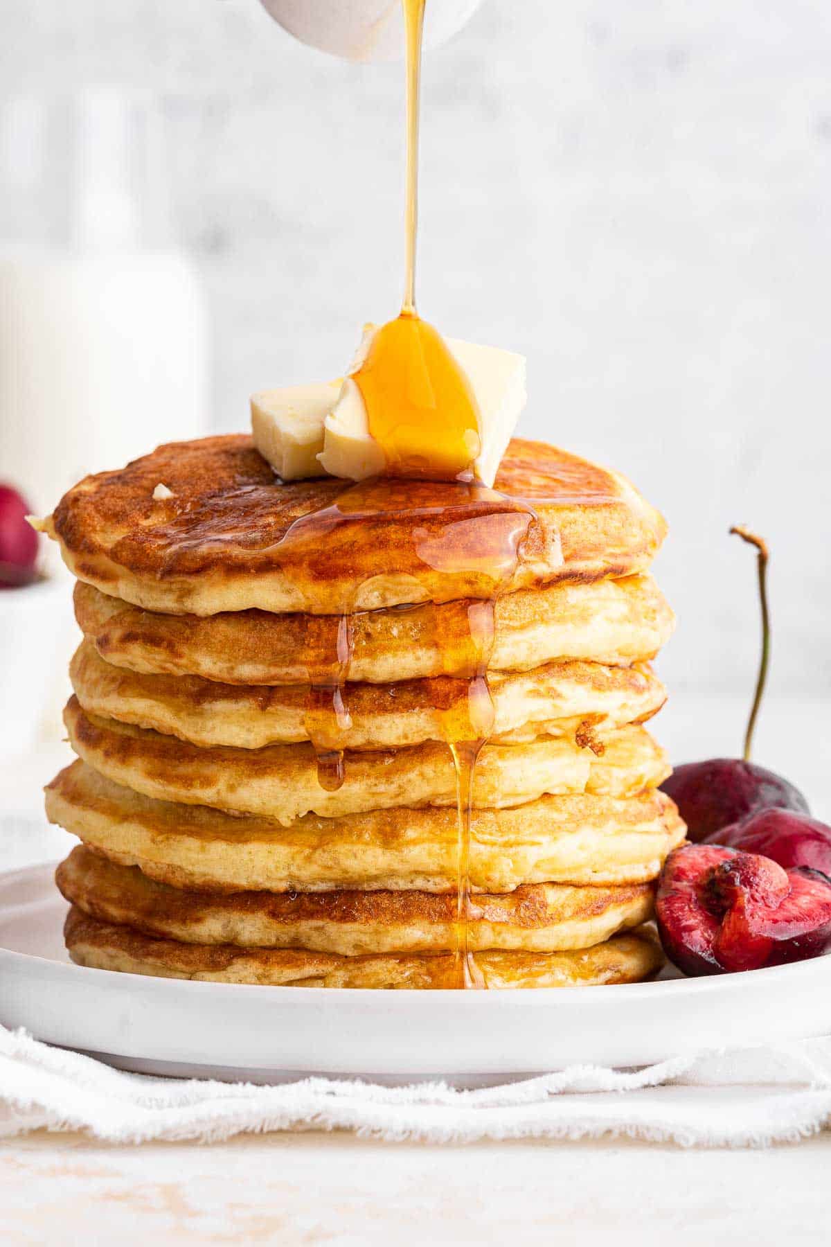 Stack of homemade pancake recipe with butter, syrup and fresh cherries on side.