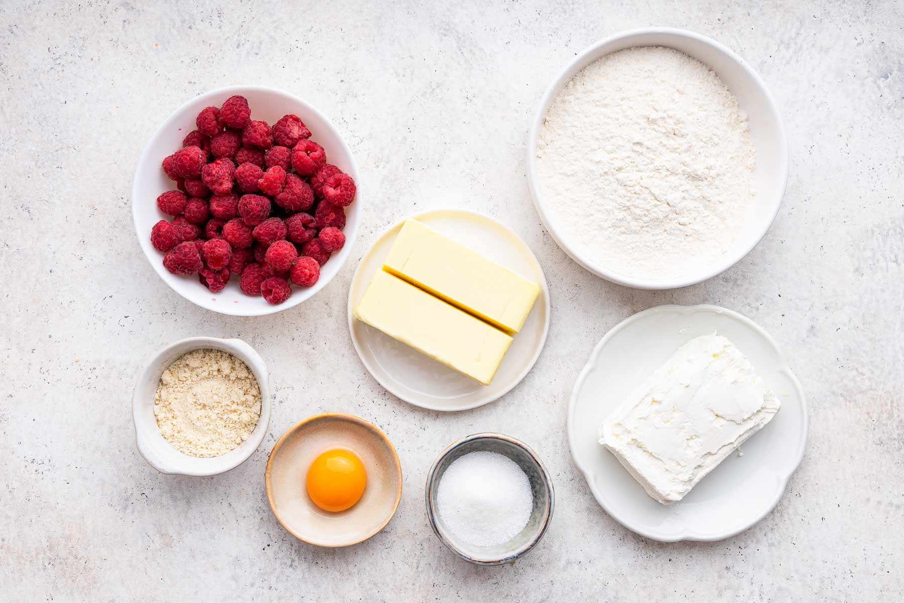 Bowl of raspberries, egg yolk, butter, sugar, and almond flour on grey surface.