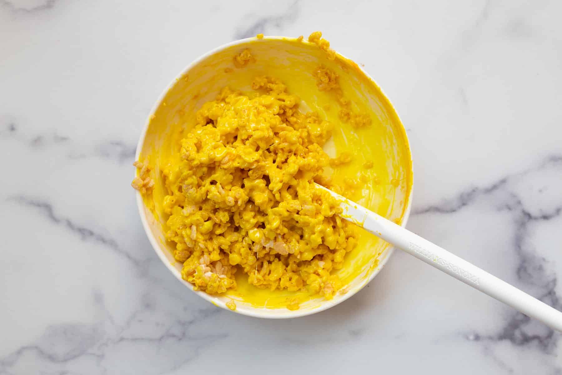 Bright yellow cereal dessert in white bowl with white spatula.