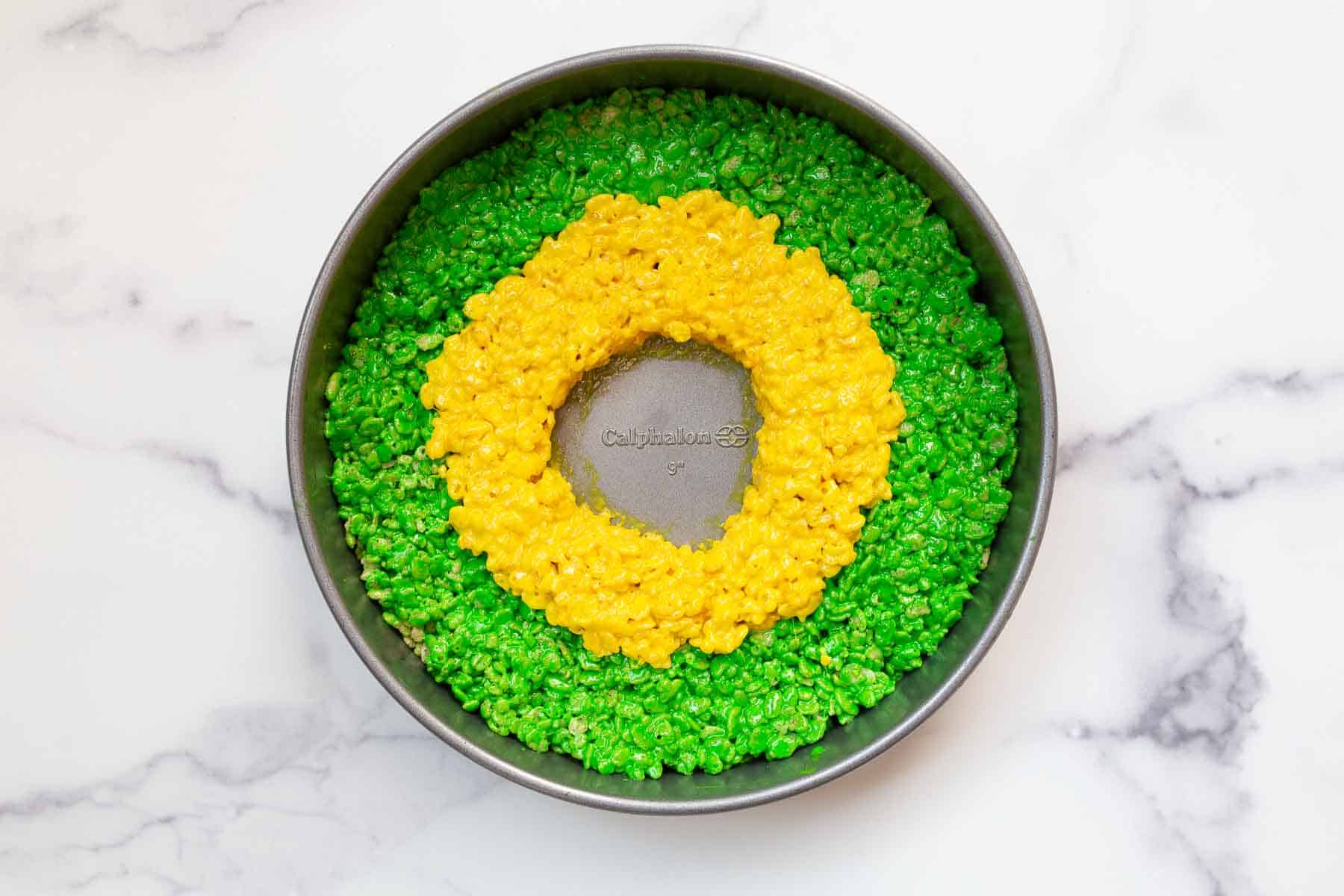 Ring of green and yellow dessert in a round cake pan on marble surface.