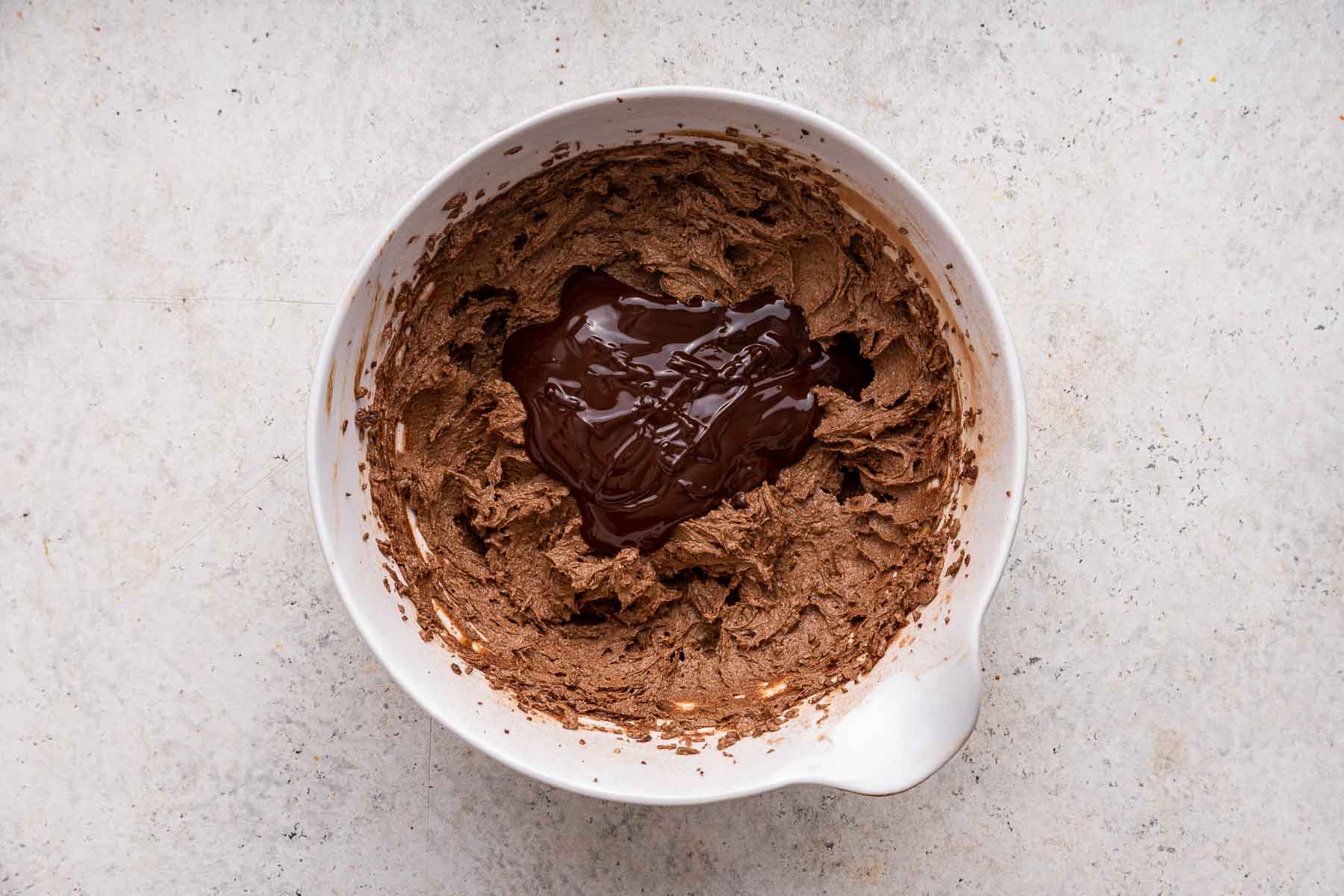 Chocolate frosting in white bowl with pile of melted chocolate poured on top.