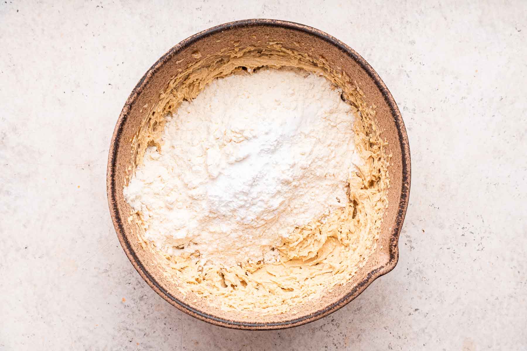 White flour sprinkled on top of yellow batter in brown mixing bowl.