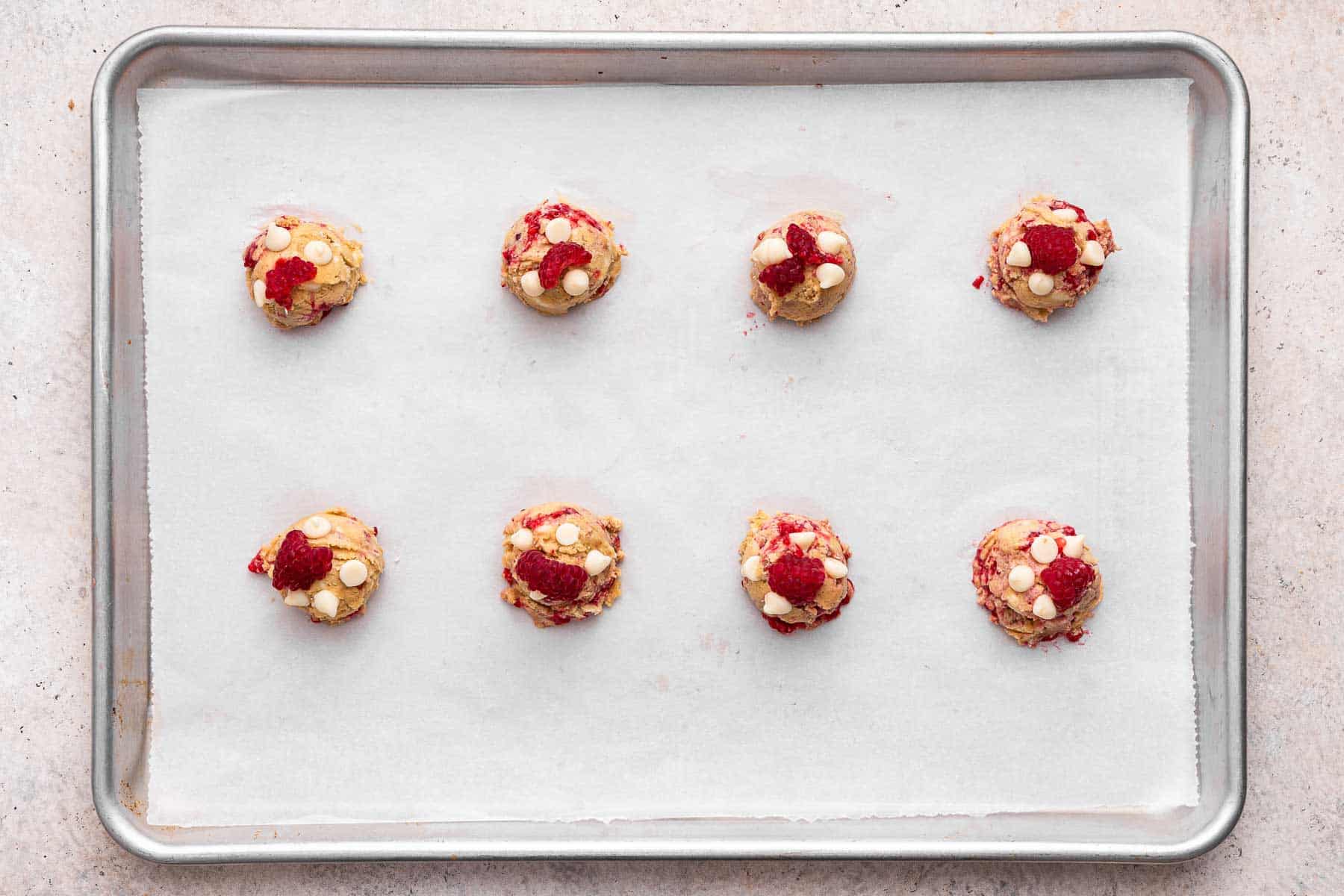 Eight balls of cookie dough with fresh raspberries on a cookie sheet.