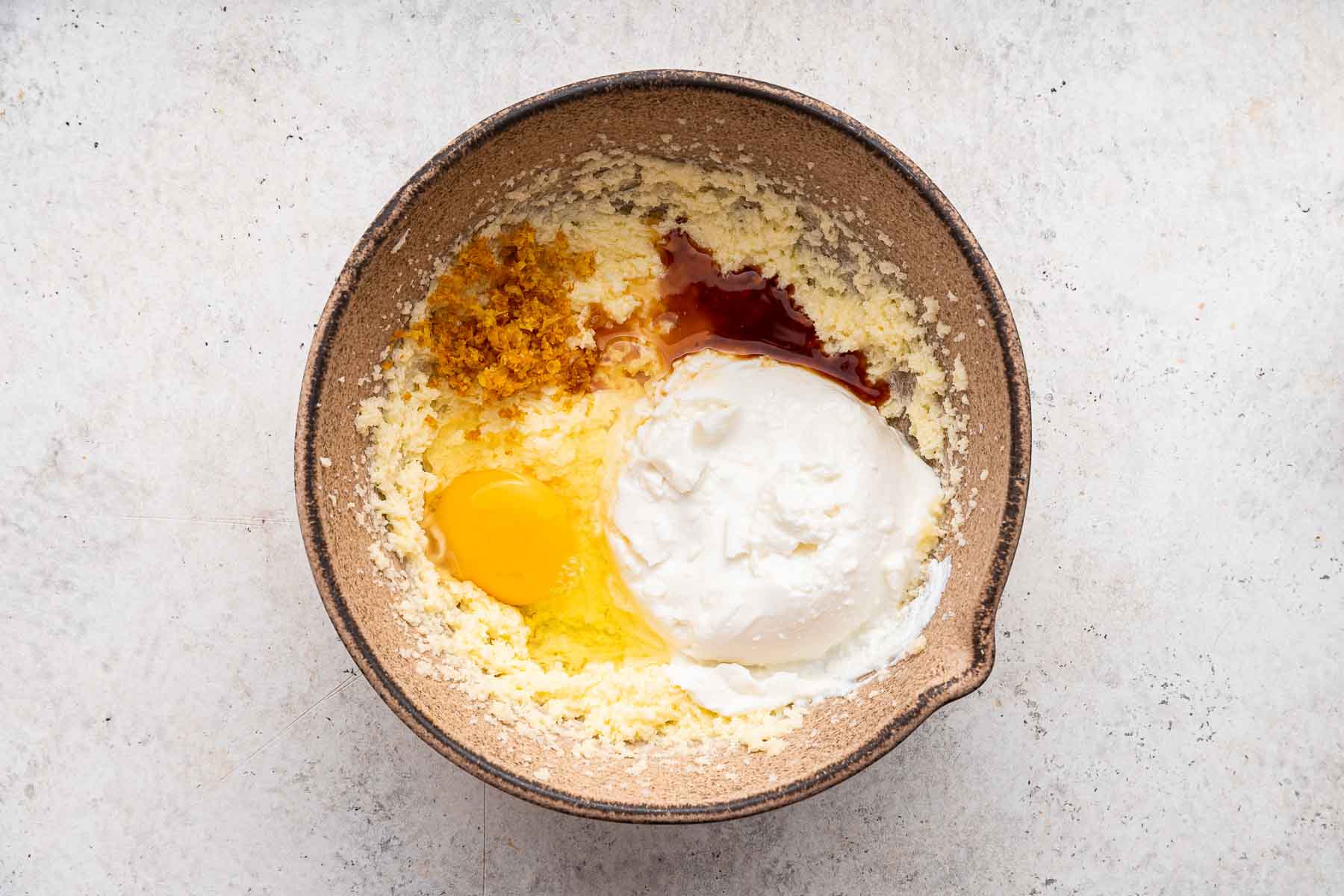Yellow batter with an egg, vanilla and lemon zest on top in brown bowl.