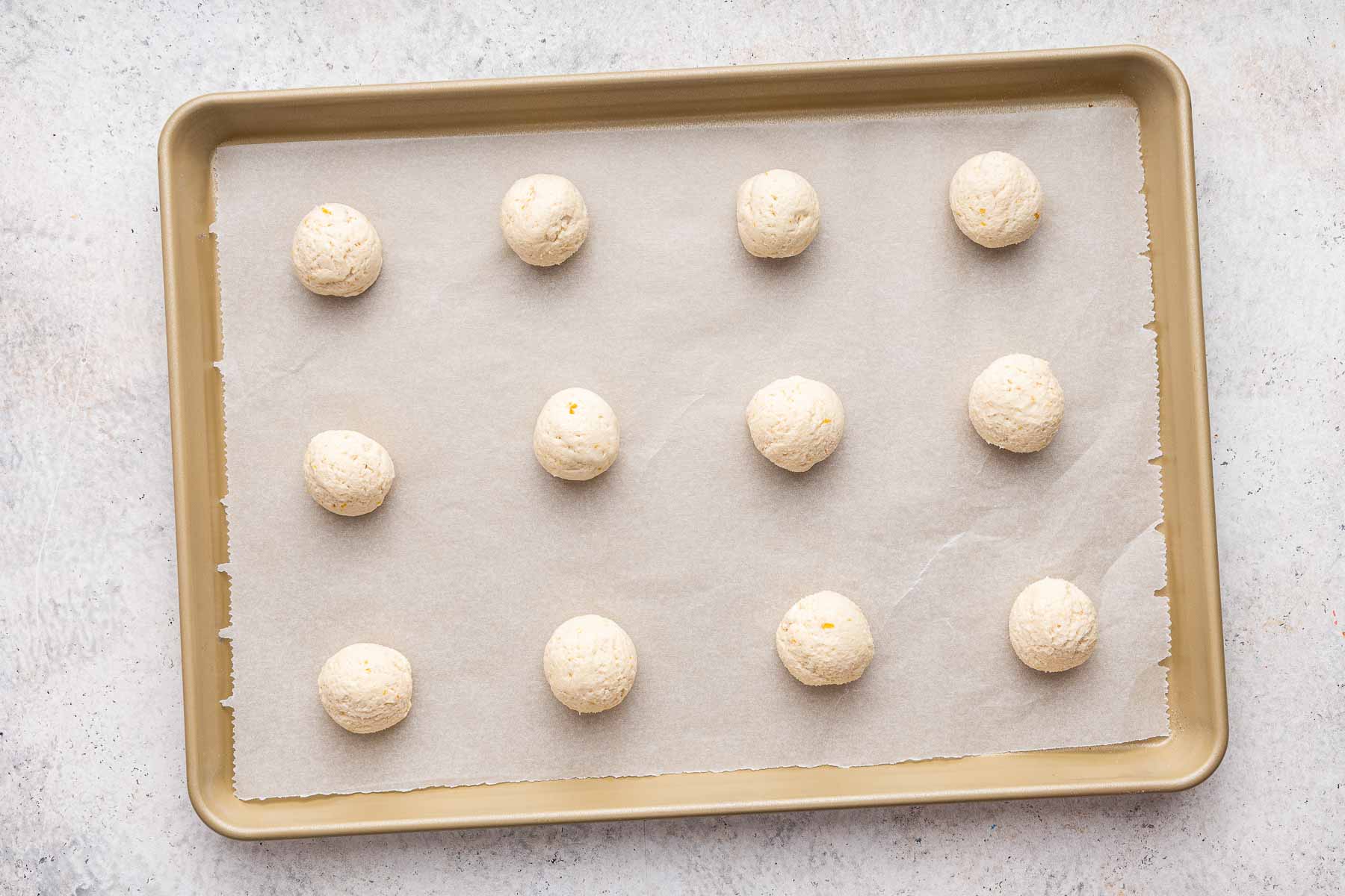 Twelve white dough balls on gold cookie sheet lined with parchment paper.