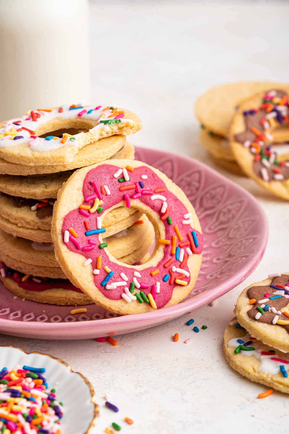 Donut cookies stacked on a pink plate with milk behind it.