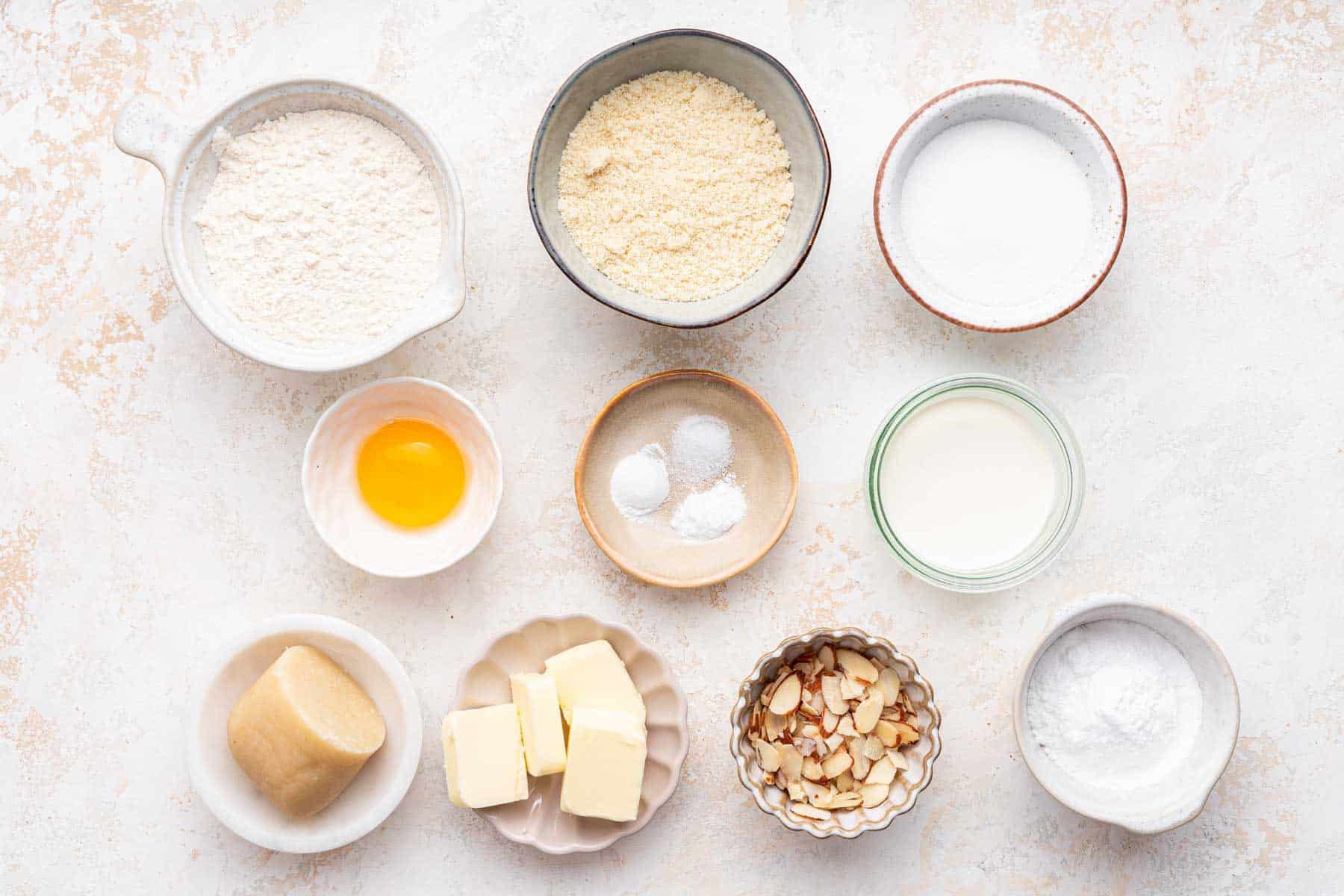 Flours, sugars and egg yolk in small bowls on white counter.