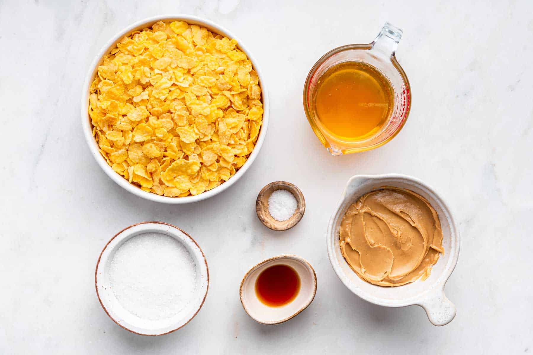 Bowl of cornflakes, sugar, honey, and peanut butter on counter.
