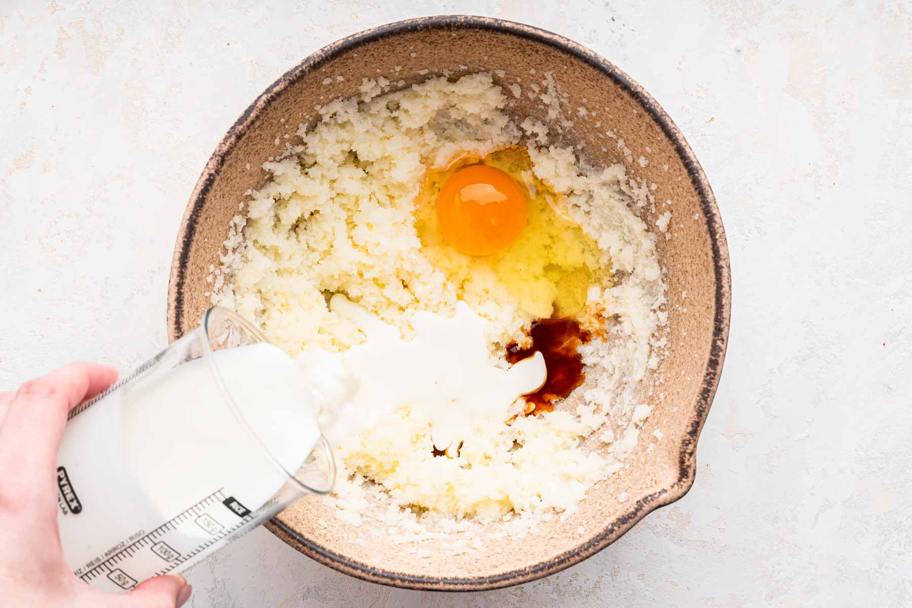 Yellow batter with egg and vanilla, and a hand pouring cream into the bowl.